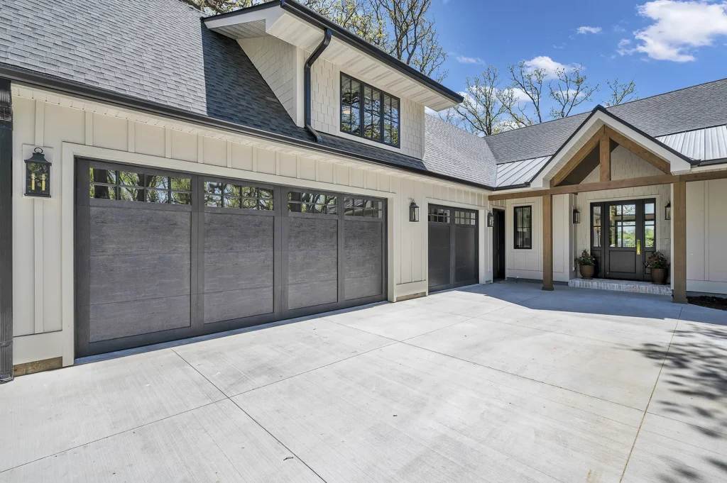 Modern Giovanti Home exterior with a large concrete driveway, three-car garage with dark doors, board and batten siding, and a covered front entry featuring potted plants and wood accents under a blue sky.